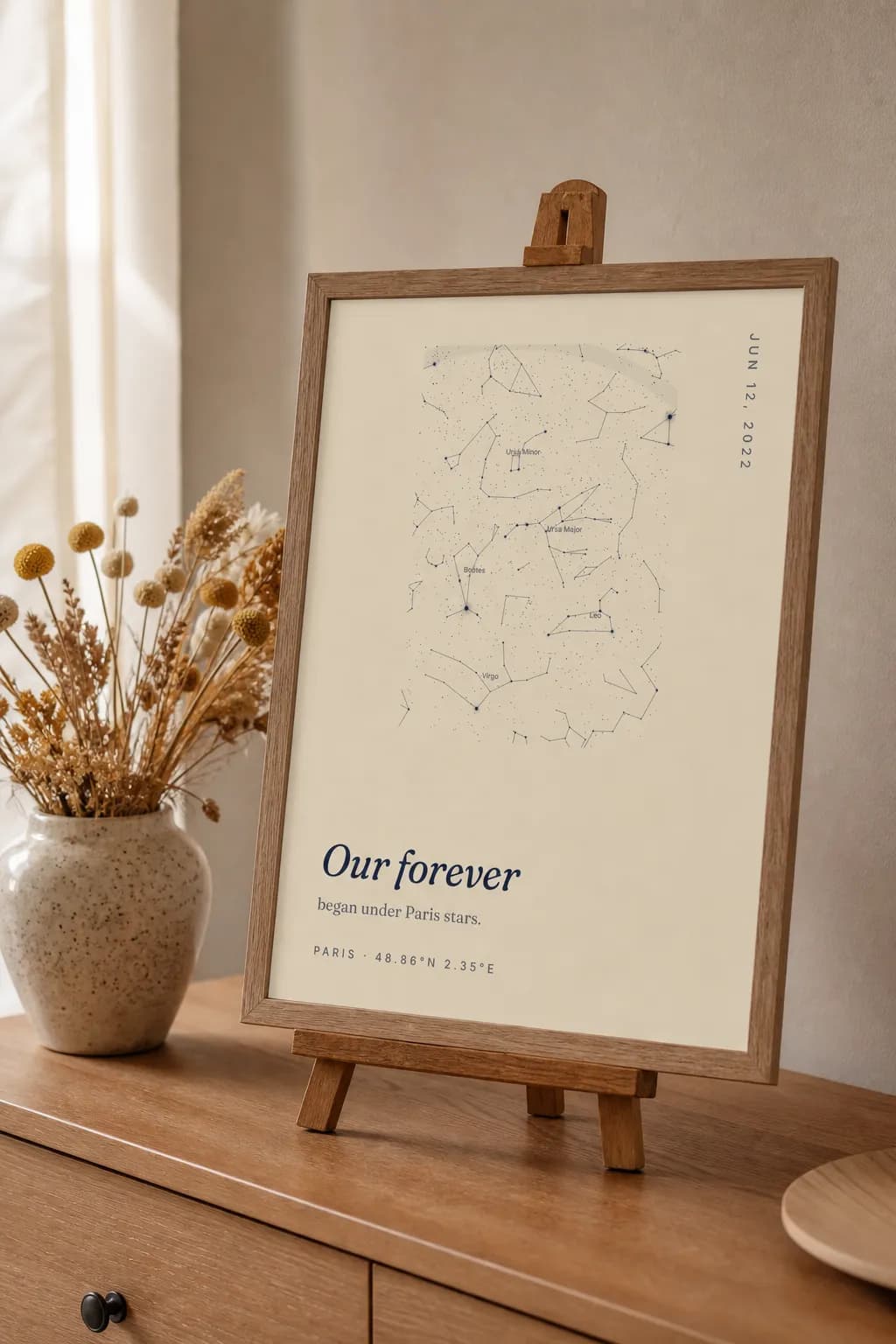 A framed SkyWhen star map print on a tabletop easel beside dried flowers